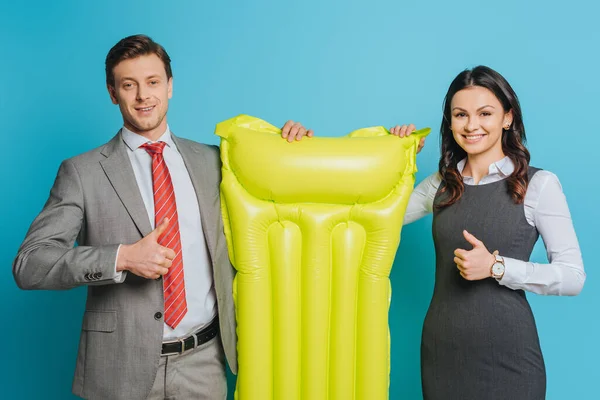 Two happy businesspeople holding yellow inflatable mattress and showing thumbs up on blue background — Stock Photo