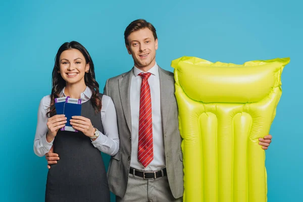 Sonriente hombre de negocios sosteniendo colchón inflable y abrazando a la mujer de negocios mostrando pasaportes y boletos aéreos aislados en azul - foto de stock