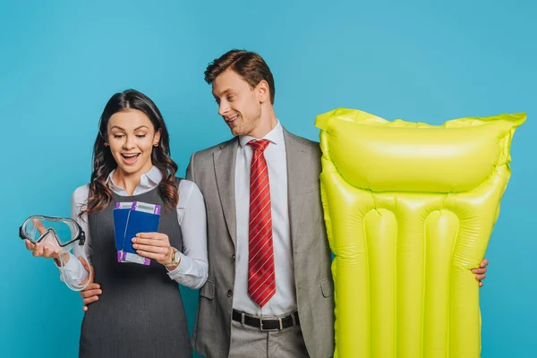 Excited businesswoman looking at passports and air tickets while holding diving mask near businessman with inflatable mattress isolated on blue — Stock Photo