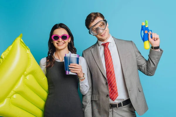 Cheerful businesswoman with inflatable mattress, passports and air tickets near businessman in diving mask holding water gun isolated on blue — Stock Photo