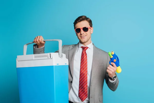 Happy businessman in sunglasses holding portable fridge and water gun on blue background — Stock Photo