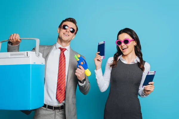Aufgeregte Geschäftsfrau mit Pässen und Flugtickets in der Nähe Geschäftsmann mit tragbarem Kühlschrank und Wasserpistole auf blauem Hintergrund — Stockfoto