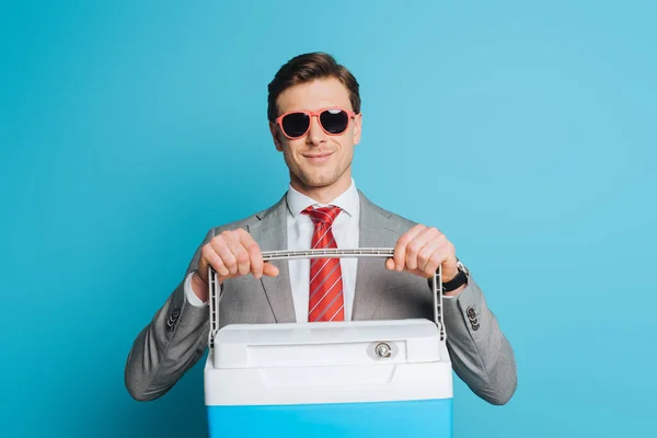 Щасливий бізнесмен в сонцезахисних окулярах тримає портативний холодильник на синьому фоні — стокове фото