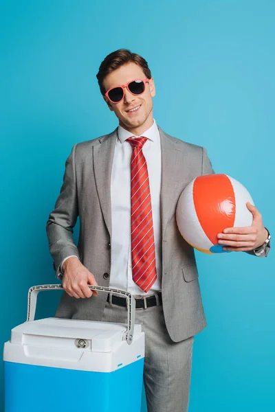 Smiling businessman in sunglasses holding portable fridge and inflatable ball on blue background — Stock Photo