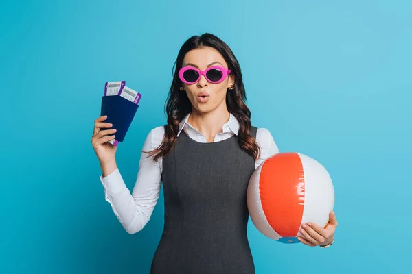 Empresária surpreendida em óculos de sol segurando bola inflável, passaportes e passagens aéreas no fundo azul — Fotografia de Stock