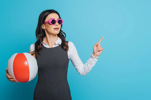 Empresária surpreso em óculos de sol segurando bola inflável, olhando para longe e apontando com o dedo no fundo azul — Fotografia de Stock