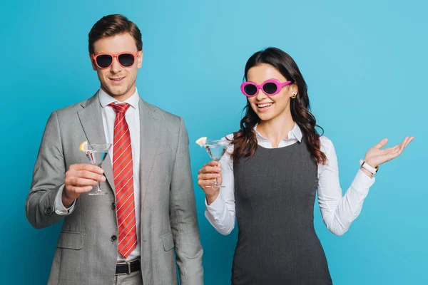 Alegre hombre de negocios y mujer de negocios en gafas de sol celebrando vacaciones con copas de cóctel sobre fondo azul - foto de stock