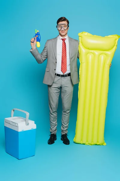 Joven hombre de negocios en máscara de buceo sosteniendo colchón inflable y pistola de agua mientras está de pie cerca de nevera portátil sobre fondo azul - foto de stock