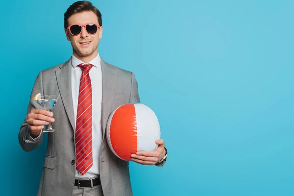Happy businessman in sunglasses holding glass of cocktail and inflatable ball on blue background — Stock Photo