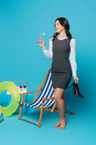 Mujer de negocios feliz sosteniendo zapatos de tacón alto y vaso de cóctel cerca de la silla de cubierta, nevera portátil y anillo inflable sobre fondo azul - foto de stock