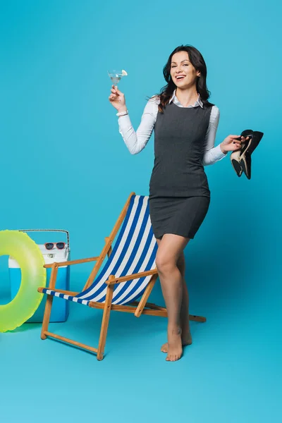 Весела бізнес-леді тримає взуття на високих підборах і склянку коктейлю біля стільця палуби, портативний холодильник і надувне кільце на синьому фоні — стокове фото