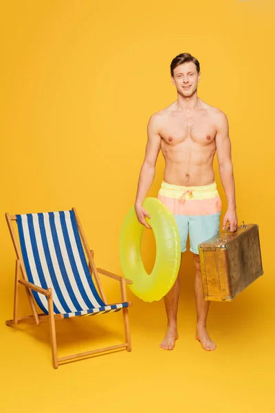 Shirtless man in shirts holding inflatable ring and vintage suitcase while standing near deck chair on yellow background — Stock Photo