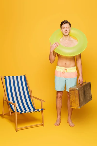 Hombre sin camisa en pantalones cortos con maleta vintage y anillo de natación de pie cerca de la silla de cubierta sobre fondo amarillo - foto de stock
