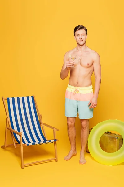 Hombre sin camisa en pantalones cortos sosteniendo vaso de cóctel cerca de la silla de cubierta, anillo de natación y maleta vintage sobre fondo amarillo - foto de stock
