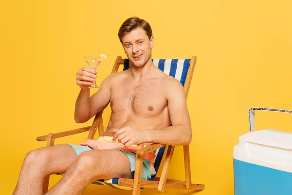 Homem sem camisa feliz sentado em cadeira de praia perto de geladeira portátil e segurando um copo de coquetel no fundo amarelo — Fotografia de Stock