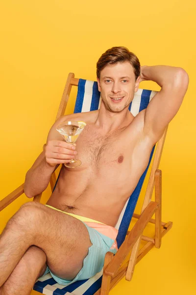 Красивий без сорочки чоловік сидить у стільці і тримає склянку коктейлю ізольовано на жовтому — стокове фото