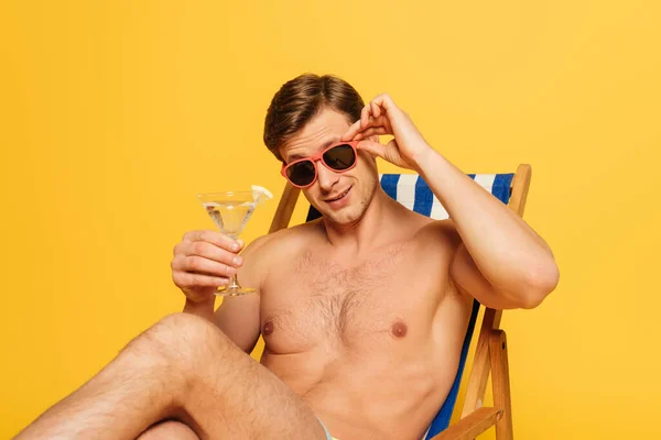 Schöner Mann mit Sonnenbrille, während er im Liegestuhl sitzt, in die Kamera blickt und ein Glas Cocktail isoliert auf gelb hält — Stockfoto