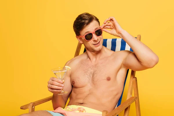 Hombre guapo tocando gafas de sol mientras está sentado en la silla de cubierta, mirando hacia otro lado y sosteniendo vaso de cóctel aislado en amarillo - foto de stock