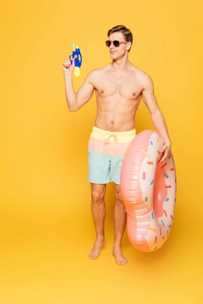 Людина в шортах і сонцезахисних окулярах тримає плавати кільце і водяний пістолет, дивлячись на жовтий фон — стокове фото