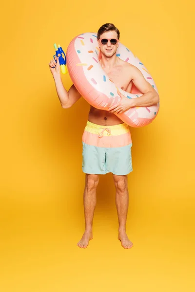 Hombre en pantalones cortos y gafas de sol sosteniendo anillo de natación y pistola de agua sobre fondo amarillo - foto de stock