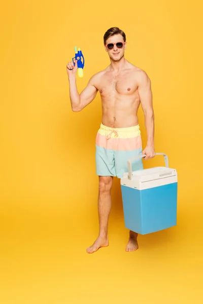 Shirtless man in shorts and sunglasses holding portable fridge and water gun on yellow background — Stock Photo