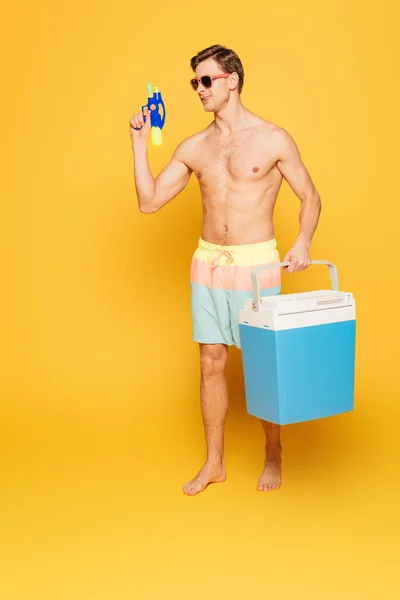 Handsome man in shorts holding portable fridge and water gun on yellow background — Stock Photo
