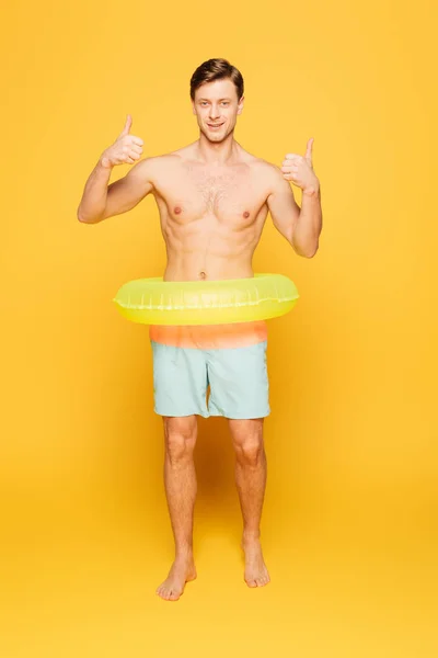 Sonriente hombre en el anillo de natación mostrando el pulgar hacia arriba mientras mira a la cámara en el fondo amarillo - foto de stock