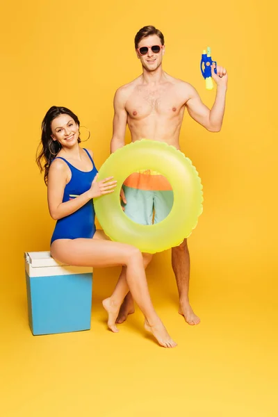 Cheerful woman sitting on portable fridge with swim ring near shirtless man holding water gun and on yellow background — Stock Photo