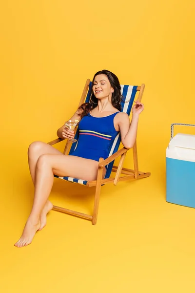 Mulher feliz com copo de coquetel sentado em cadeira de praia com olhos fechados perto de geladeira portátil no fundo amarelo — Fotografia de Stock