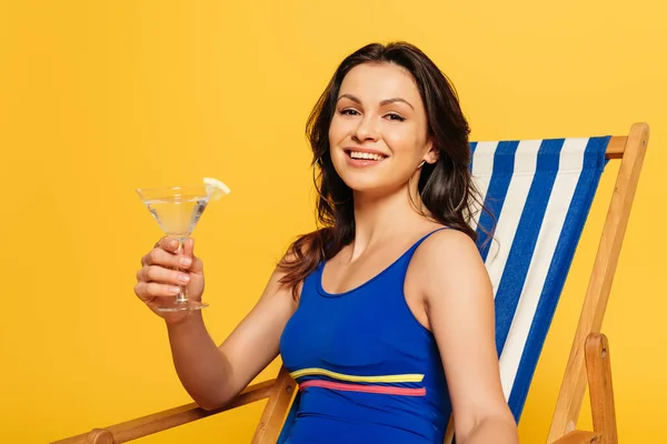 Fröhliche Frau im blauen Badeanzug, die im Liegestuhl mit einem Glas Cocktail sitzt und isoliert auf gelb in die Kamera schaut — Stockfoto