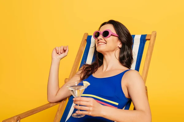 Mujer feliz en traje de baño y gafas de sol descansando en la silla de cubierta mientras sostiene el vaso de cóctel aislado en amarillo - foto de stock