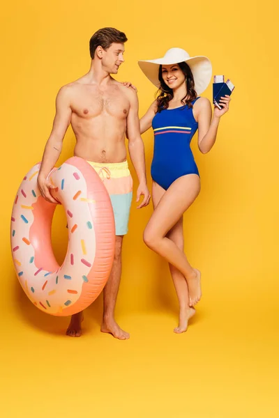 Happy woman in swimsuit holding passports and air tickets while touching shoulder of shirtless man with inflatable ring on yellow background — Stock Photo