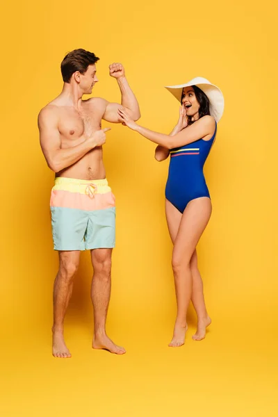 Shocked woman in swimsuit touching biceps of handsome shirtless man on yellow background — Stock Photo