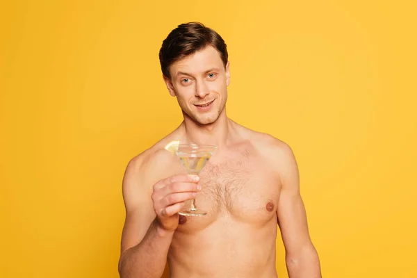 Schöner hemdloser Mann, der in die Kamera schaut, während er ein Glas Cocktail auf gelbem Grund hält — Stockfoto