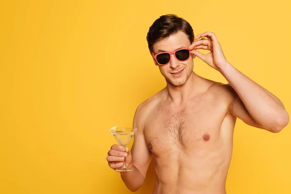 Hombre sin camisa tocando gafas de sol mientras sostiene el vaso de cóctel sobre fondo amarillo - foto de stock