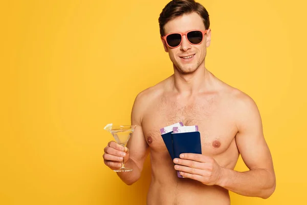 Shirtless man in sunglasses holding glass of cocktail, passports and air tickets on yellow background — Stock Photo