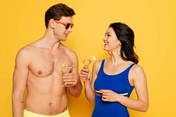 Feliz pareja en gafas de sol hablando mientras sostiene vasos de cóctel sobre fondo amarillo - foto de stock