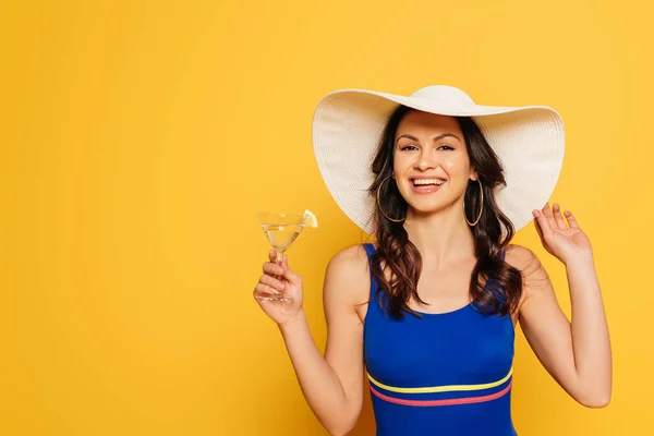 Улыбающаяся женщина в синем купальнике и шляпе, держащая стакан коктейля и смотрящая на камеру, изолированную на желтом — стоковое фото