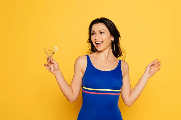 Веселая женщина в синем купальнике держит стакан коктейля и смотрит в сторону изолированной на желтый — стоковое фото
