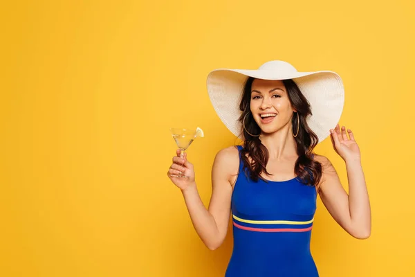 Щаслива жінка в купальнику, торкаючись сонячного капелюха, тримаючи склянку коктейлю ізольовано на жовтому — стокове фото