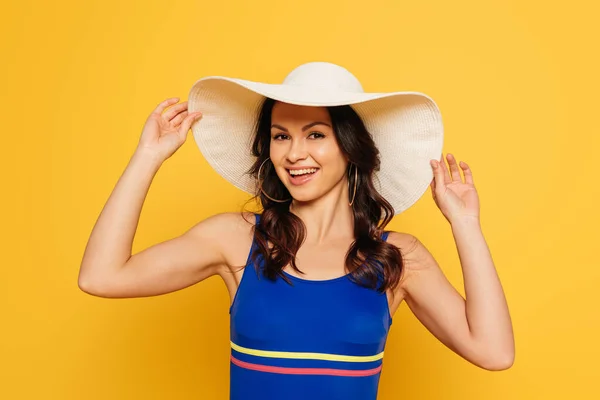 Femme heureuse en maillot de bain touchant chapeau de soleil isolé sur jaune — Photo de stock