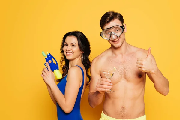 Весёлый мужчина в водолазной маске со стаканом коктейля показывает большой палец вверх рядом с улыбающейся женщиной с водяным пистолетом на жёлтом фоне — стоковое фото