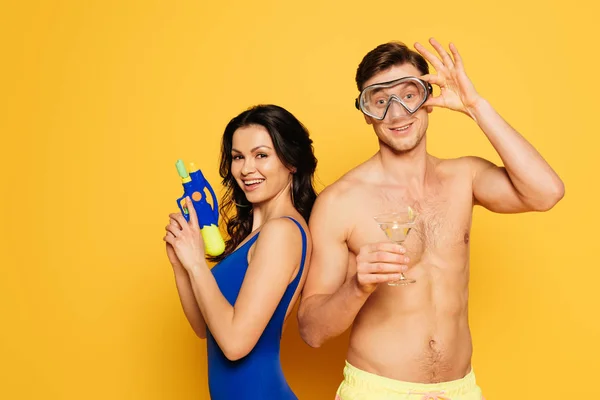 Homme gai masque de plongée tenant verre de cocktail près femme heureuse avec pistolet à eau sur fond jaune — Photo de stock