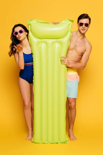 Attractive woman blowing air kiss and handsome shirtless man near inflatable mattress on yellow background — Stock Photo