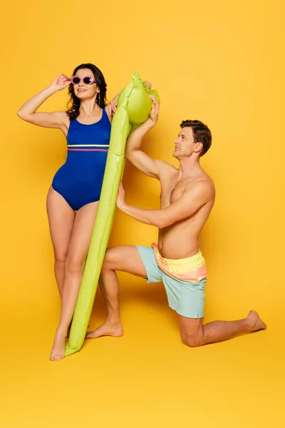 Séduisante femme en maillot de bain bleu touchant lunettes et homme torse nu debout sur le genou près du matelas gonflable sur fond jaune — Photo de stock