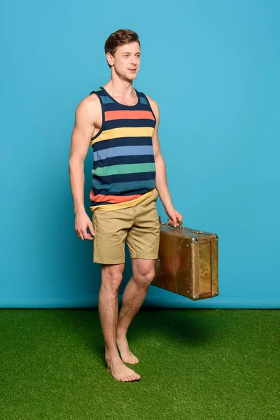 Handsome man in striped singlet and shorts holding vintage suitcase on blue background — Stock Photo