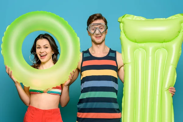 Smiling man in diving mask holding inflatable mattress near happy woman with swim ring on blue background — Stock Photo