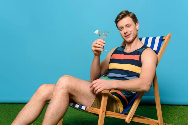 Bel homme souriant tenant un verre de cocktail assis sur une chaise longue et regardant la caméra sur fond bleu — Photo de stock