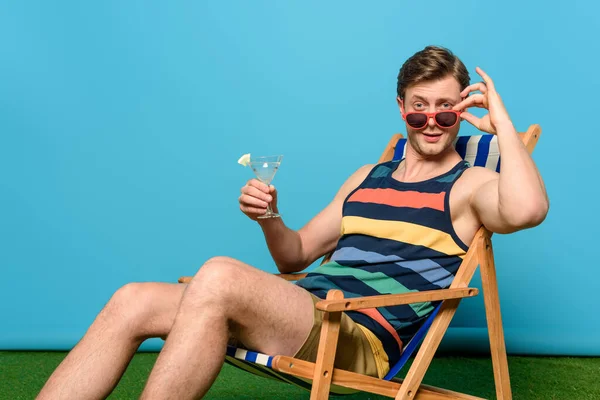 Gutaussehend mit Cocktailglas, Brille anfassen und in die Kamera schauen, während er im Liegestuhl auf blauem Hintergrund sitzt — Stockfoto