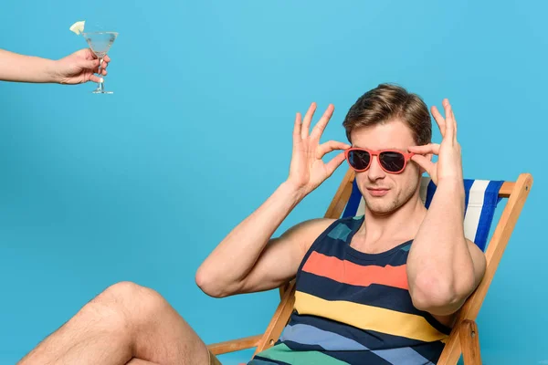 Ausgeschnittene Ansicht einer Frau, die einem Mann, der im Liegestuhl sitzt und Sonnenbrille auf blauem Hintergrund berührt, ein Glas Cocktail gibt — Stockfoto